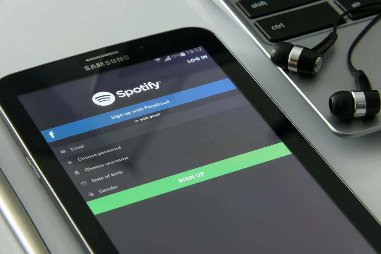 A tablet displays the Spotify sign-up screen beside a laptop and earphones. The scene conveys a tech-savvy, music-focused atmosphere.