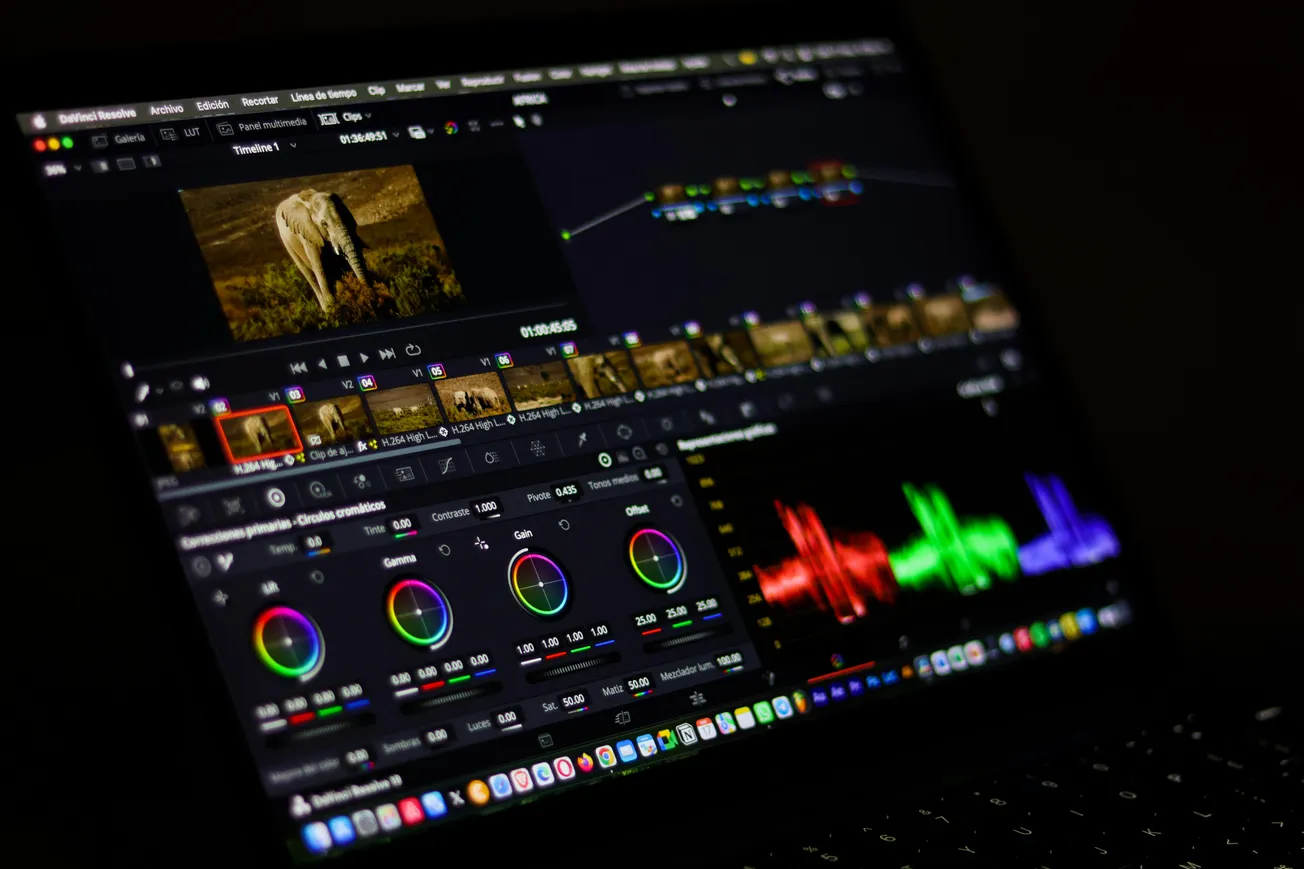 Laptop screen showing video editing software with color correction tools, a timeline of clips, and color waveforms. The scene depicts dynamic editing in progress.