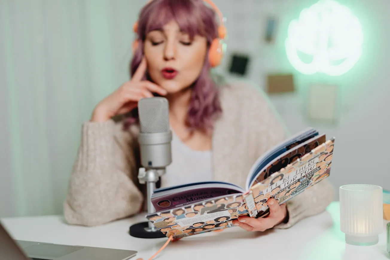 Purple-haired woman reading aloud into a microphone, wearing orange headphones. She sits in a cozy room, setting a focused, creative tone.