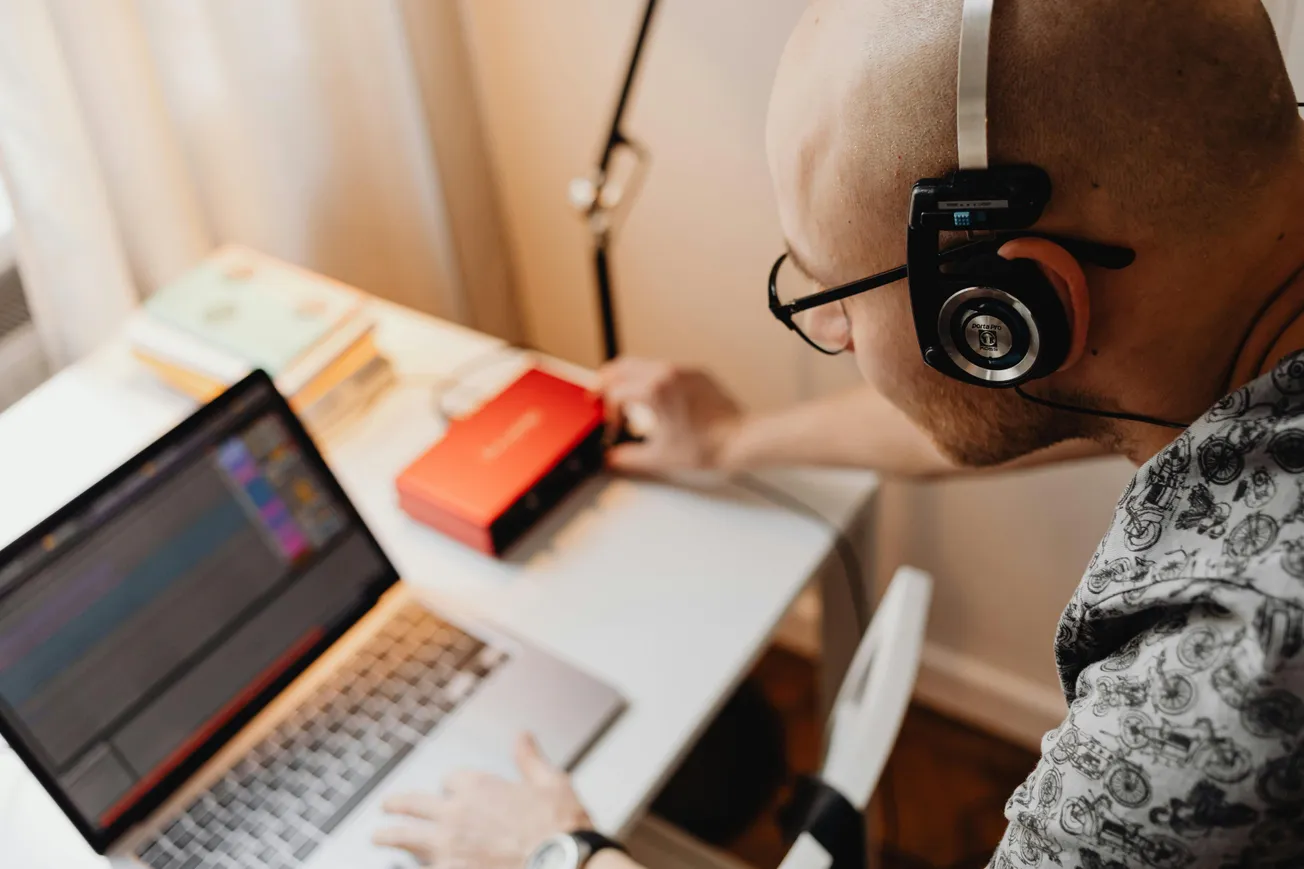 A bald person wearing headphones and glasses works on audio editing on a laptop. A red audio interface and books are on the desk, evoking a focused, creative atmosphere.