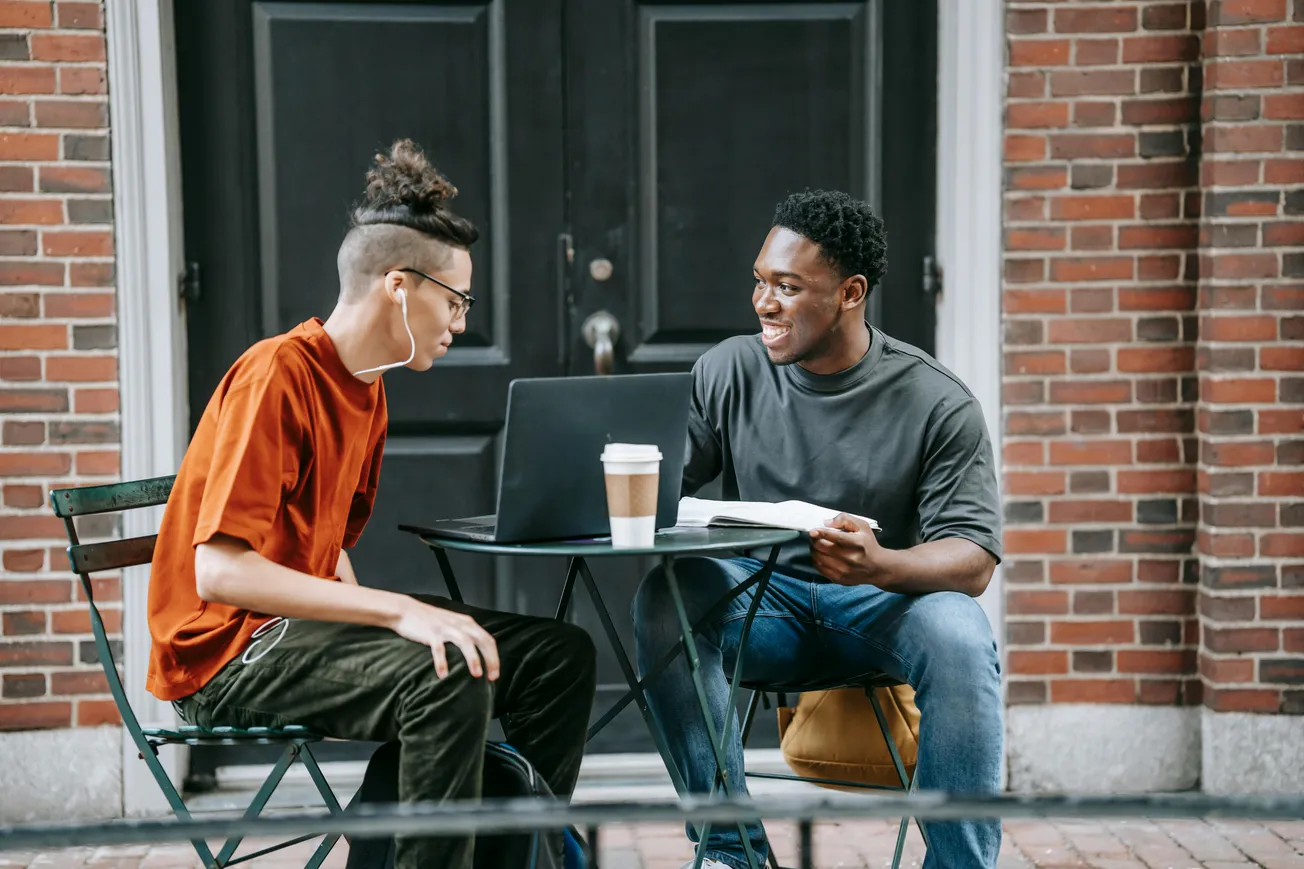 Two people sit outside a brick building at a small table. One is using a laptop, and the other holds a book. A coffee cup is on the table. Both are smiling.