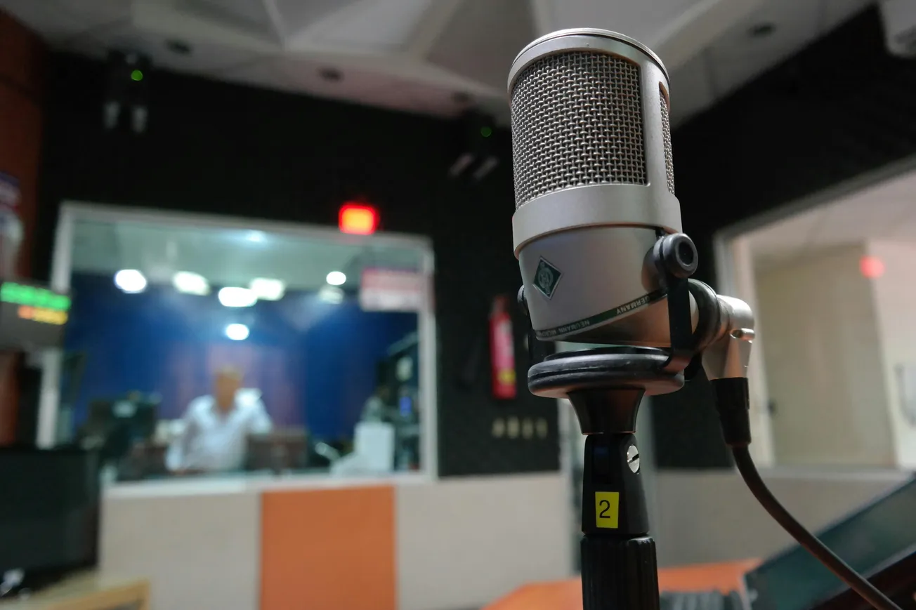 Close-up of a silver microphone in a soundproof radio studio. In the blurred background, a person sits behind a glass window, conveying a broadcast atmosphere.