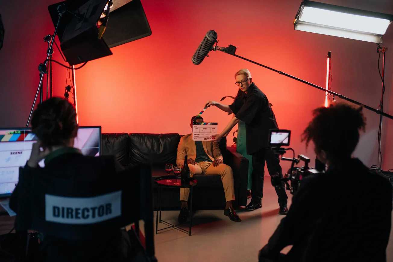 Film set with a crew in action. A person holds a clapperboard near an actor on a sofa. Warm orange lighting, with "Director" chair and camera equipment.